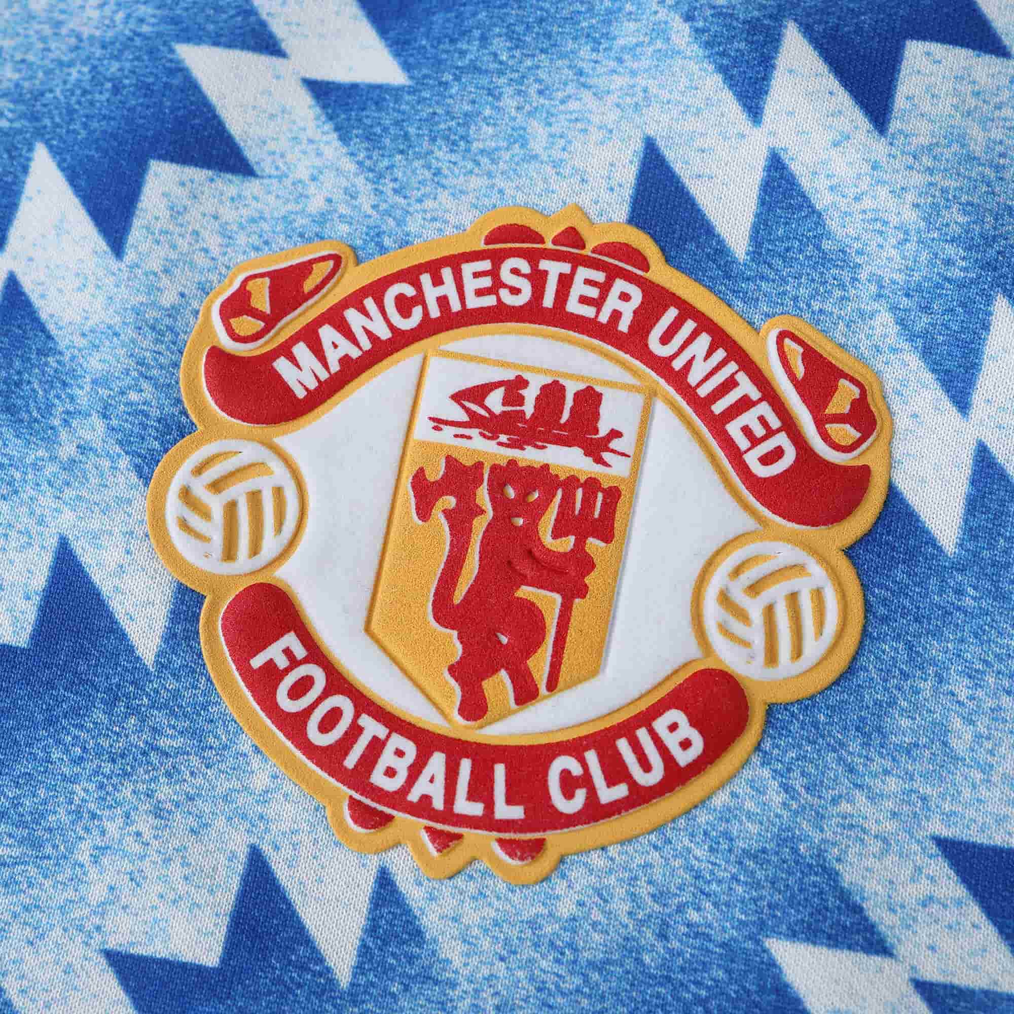 A close-up of a Manchester United Football Club crest patch on a blue and white zigzag patterned fabric background.