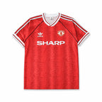 A red short-sleeve Manchester United soccer jersey, featuring white shoulder stripes, a white V-neck collar, the white logo, the Manchester United crest, and the white SHARP sponsor logo across the chest.