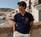A man wearing a navy blue Spain national football team jersey by Adidas, leaning against a stone wall with a coastal town background.