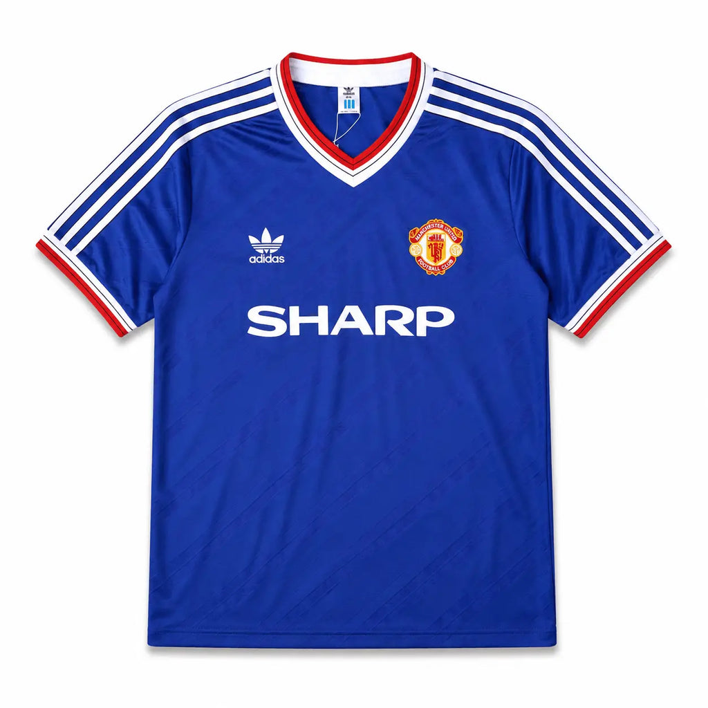 A blue Manchester United football jersey, featuring the club's crest on the left chest, the white Adidas logo on the right chest, and the sponsor 'SHARP' printed in white across the center. The jersey has a V-neck with red and white trim, and white stripes on the shoulders and sleeves.