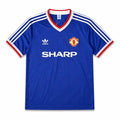 A blue Manchester United football jersey, featuring the club's crest on the left chest, the white Adidas logo on the right chest, and the sponsor 'SHARP' printed in white across the center. The jersey has a V-neck with red and white trim, and white stripes on the shoulders and sleeves.