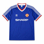 A blue Manchester United football jersey, featuring the club's crest on the left chest, the white Adidas logo on the right chest, and the sponsor 'SHARP' printed in white across the center. The jersey has a V-neck with red and white trim, and white stripes on the shoulders and sleeves.