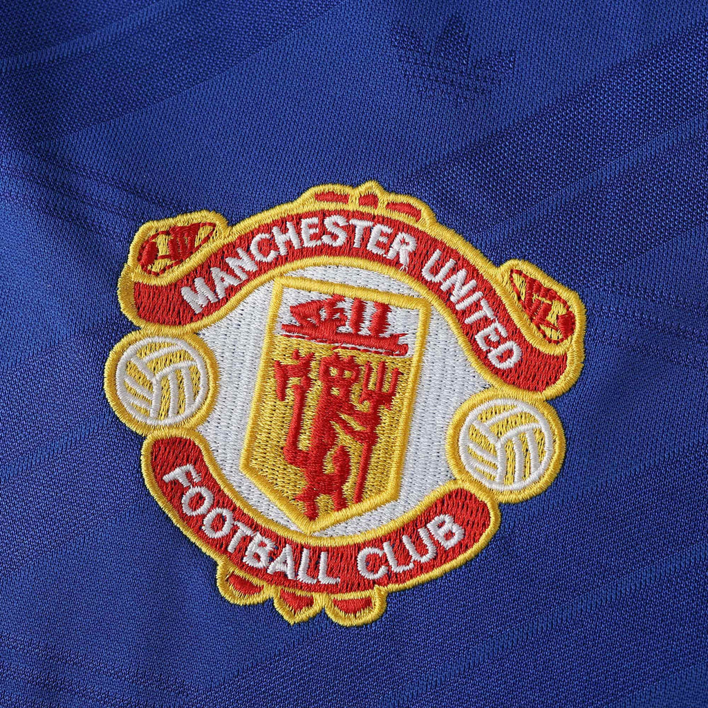 Close-up of the Manchester United Football Club embroidered crest on a royal blue textured fabric background.