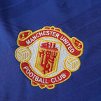 Close-up of the Manchester United Football Club embroidered crest on a royal blue textured fabric background.