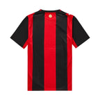 The back of a short-sleeve soccer jersey featuring bold vertical red and black stripes, with a small gold hexagonal crest centered below the collar, displayed on a solid white background.