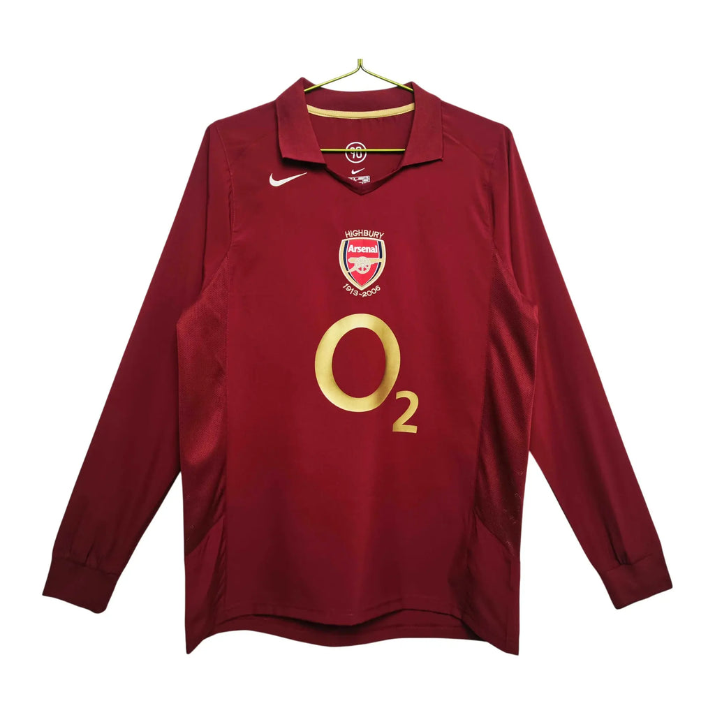 A long-sleeve Arsenal football jersey in dark red, featuring a gold O2 sponsor logo on the front, the Arsenal club crest, and a white Nike swoosh on the chest, hanging on a gold hanger against a white background.