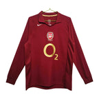 A long-sleeve Arsenal football jersey in dark red, featuring a gold O2 sponsor logo on the front, the Arsenal club crest, and a white Nike swoosh on the chest, hanging on a gold hanger against a white background.