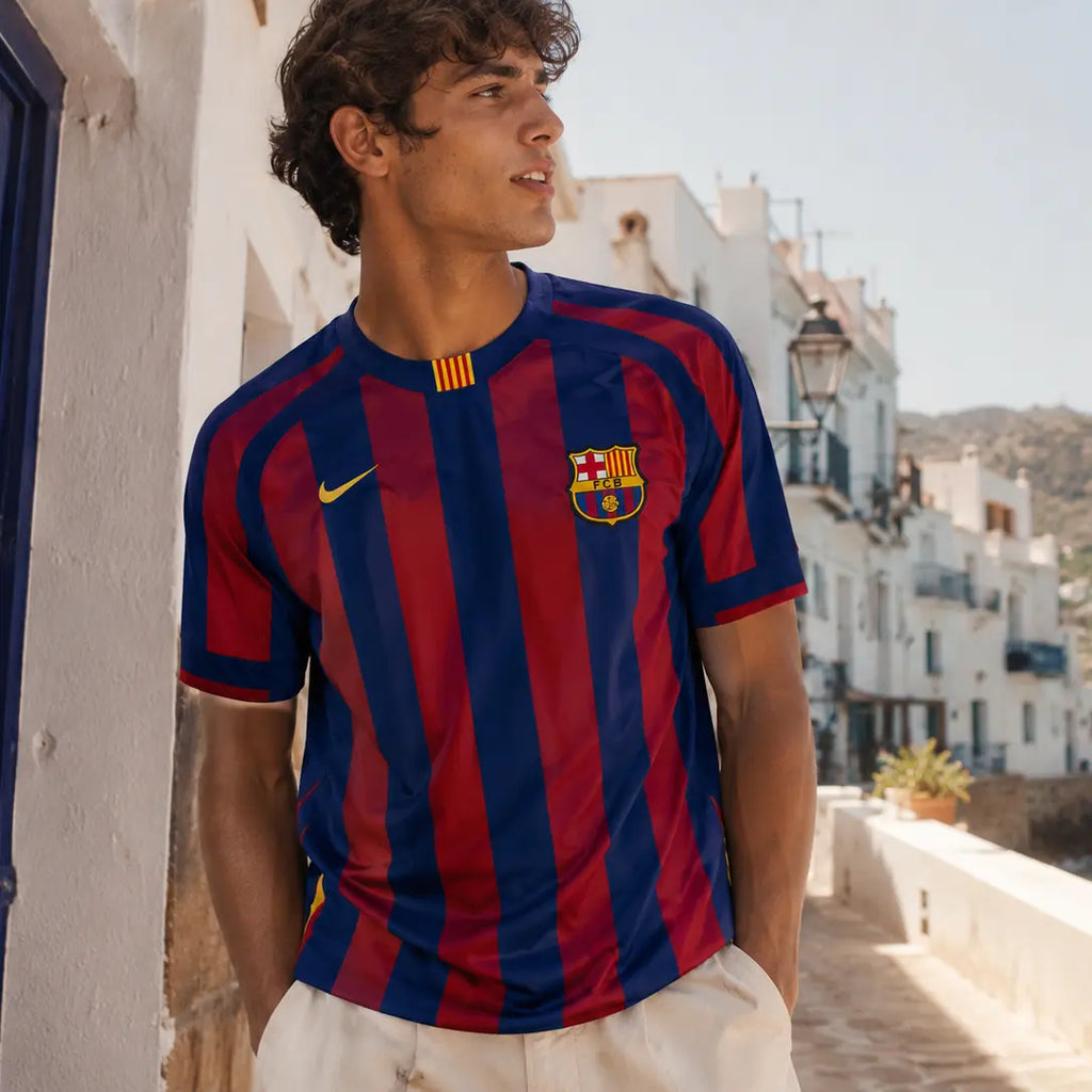 A man wearing a red and navy blue FC Barcelona soccer jersey with a yellow Nike logo, standing on a sunny coastal walkway with white buildings and blue water in the background.
