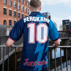 A man viewed from behind wearing a blue Arsenal football jersey with 'BERGKAMP' and the number '10' on the back, standing on a bridge with a brick building in the background.