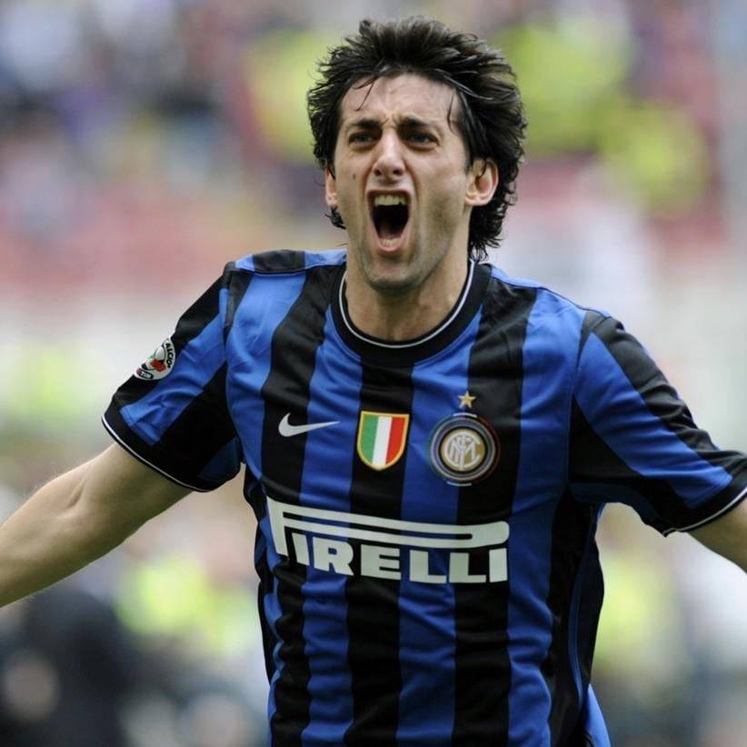A soccer player wearing an Inter Milan jersey with blue and black vertical stripes, celebrating with his arms outstretched on a stadium field.
