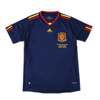 A navy blue Adidas Spain soccer jersey with red and yellow trim on the collar and sleeve cuffs, featuring the Spain national team crest and gold Adidas logo on the chest, displayed against a solid black background.