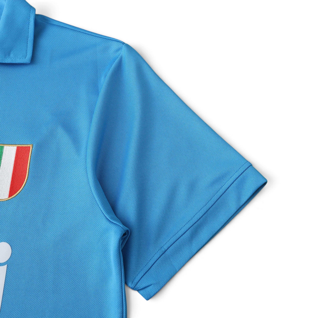 Elite Retro Kits sky blue Napoli 1987/88 home football shirt with Scudetto shield detail on a green pitch background.