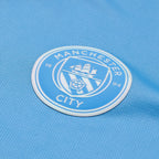 A close-up of a Manchester City soccer jersey in light blue, featuring the circular club crest patch on the chest.